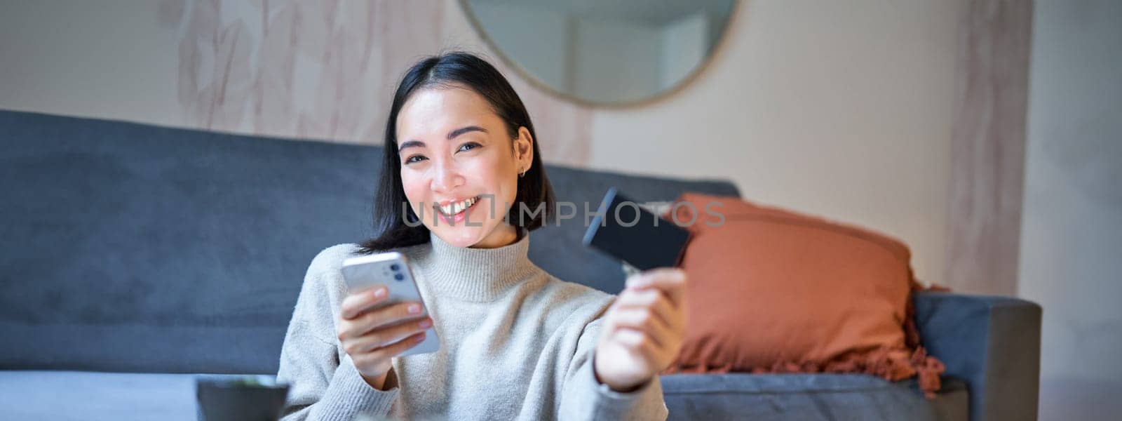Smiling asian girl with smartphone and credit card, does her shopping online, uses mobile phone to order home delivery.