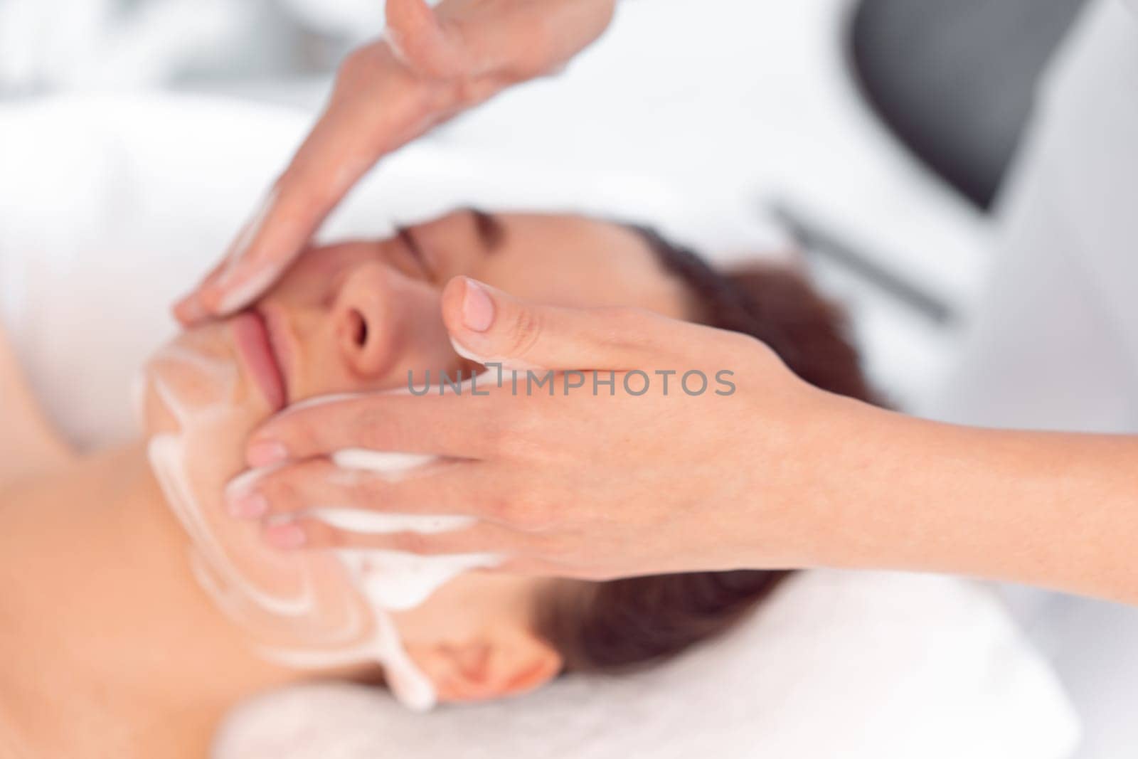 Crop anonymous cosmetologist massaging woman's face at beauty salon. From above of relaxed female customer is getting pampered in spa by beautician.