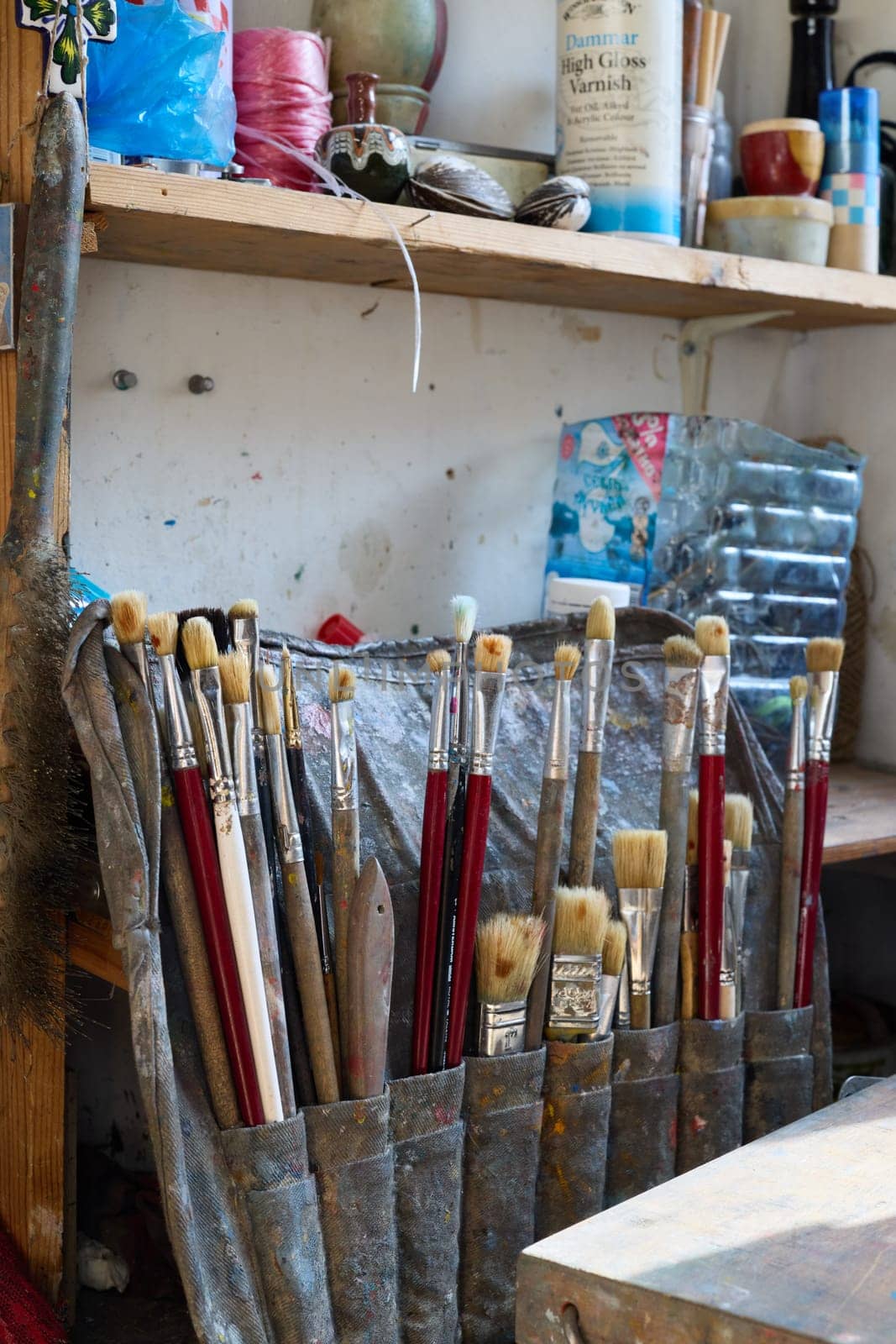 A cluttered workspace features an array of brushes, tools, and containers, illuminated by natural light from a nearby window, reflecting an artist's vibrant creativity.