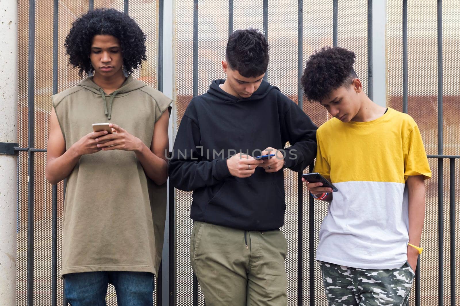 three serious multiracial teen friends browsing Internet on mobile phones standing against metal fence and looking at screen, concept of youth and technology of communication