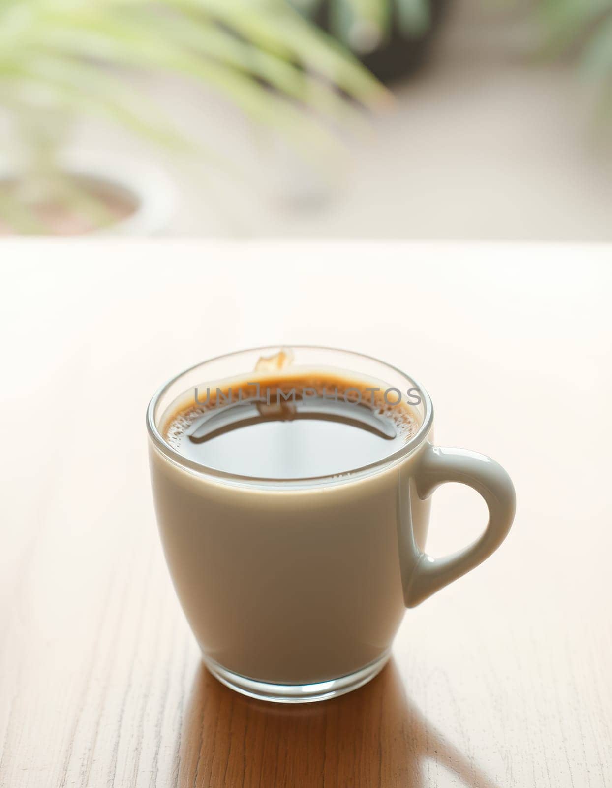 A clear glass mug filled with rich, dark coffee is placed on a light wooden table. Gentle sunlight filters through a nearby window, creating a warm atmosphere.
