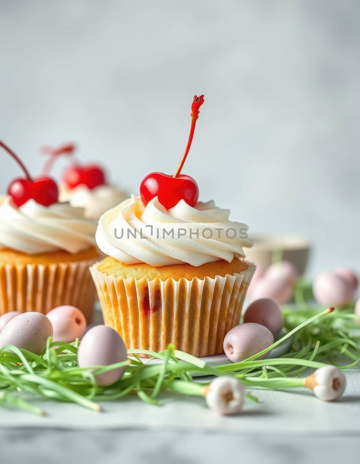 Colorful cupcakes adorned with whipped cream and cherries sit on a soft grey surface, accompanied by pastel candy accents and fresh green grass-like decor, creating a festive atmosphere.