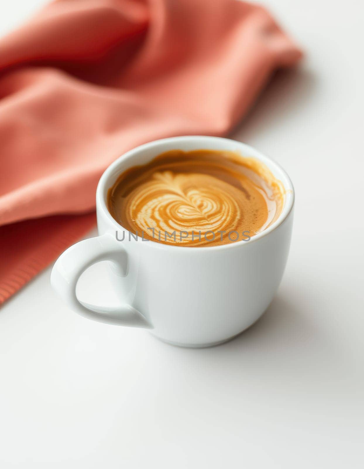 A white cup filled with creamy latte showcases beautiful swirls of latte art. A soft peach-colored cloth sits beside it, creating a cozy atmosphere.
