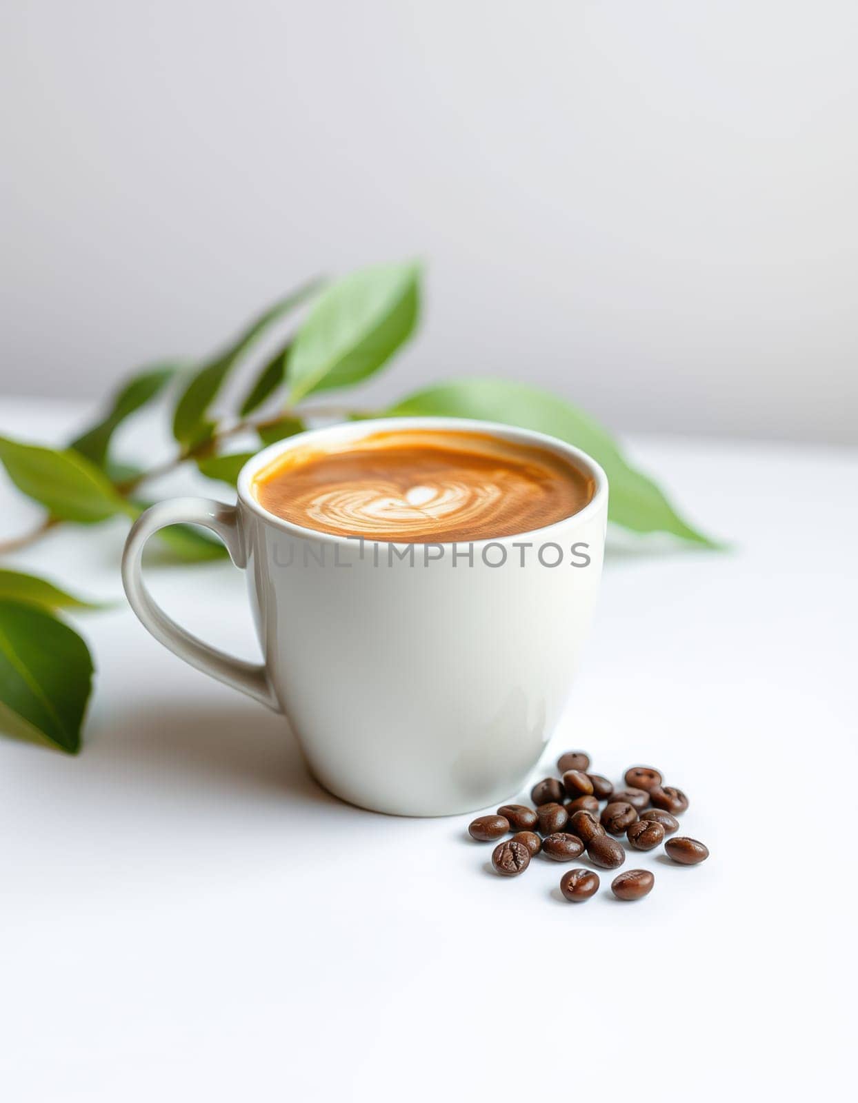 A white cup holds a perfectly crafted latte topped with intricate foam art, placed beside roasted coffee beans and fresh green leaves on a minimalist countertop.