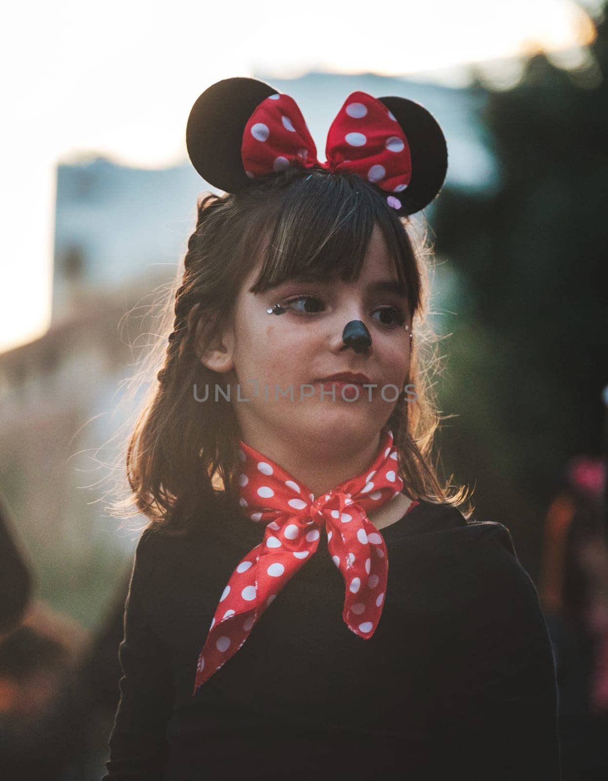 The image shows a child dressed in a costume that includes what looks like Mouse ears, commonly associated with the iconic Disney character Minnie Mouse. The child appears to have a red bow with white polka dots, which is Minnie Mouse's signature style. Additionally, there is face paint resembling a small black nose and whiskers, contributing to the mouse-themed appearance. The child is also wearing a red scarf with polka dots, matching the bow on top of the head. The background is blurred, putting the focus on the child, who seems to be looking off into the distance.