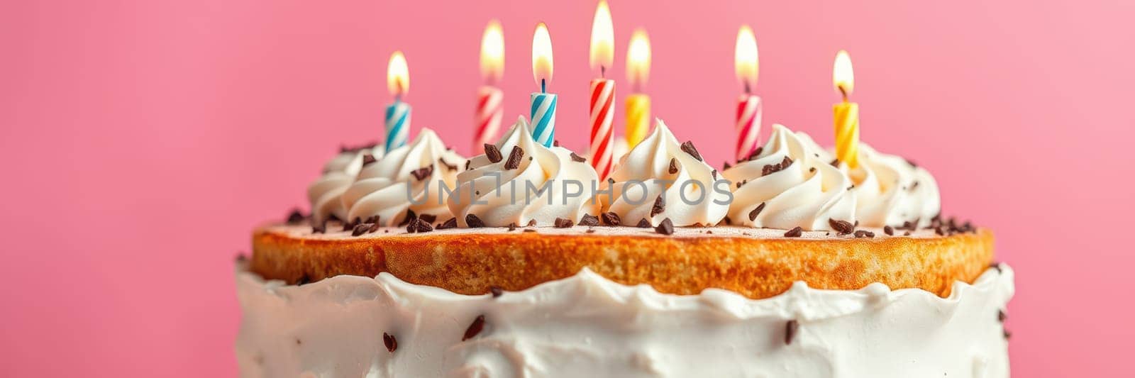 A birthday cake with creamy frosting topped with colorful sprinkles and lit candles stands against a vibrant pink backdrop. This delightful dessert is ready for a celebration.