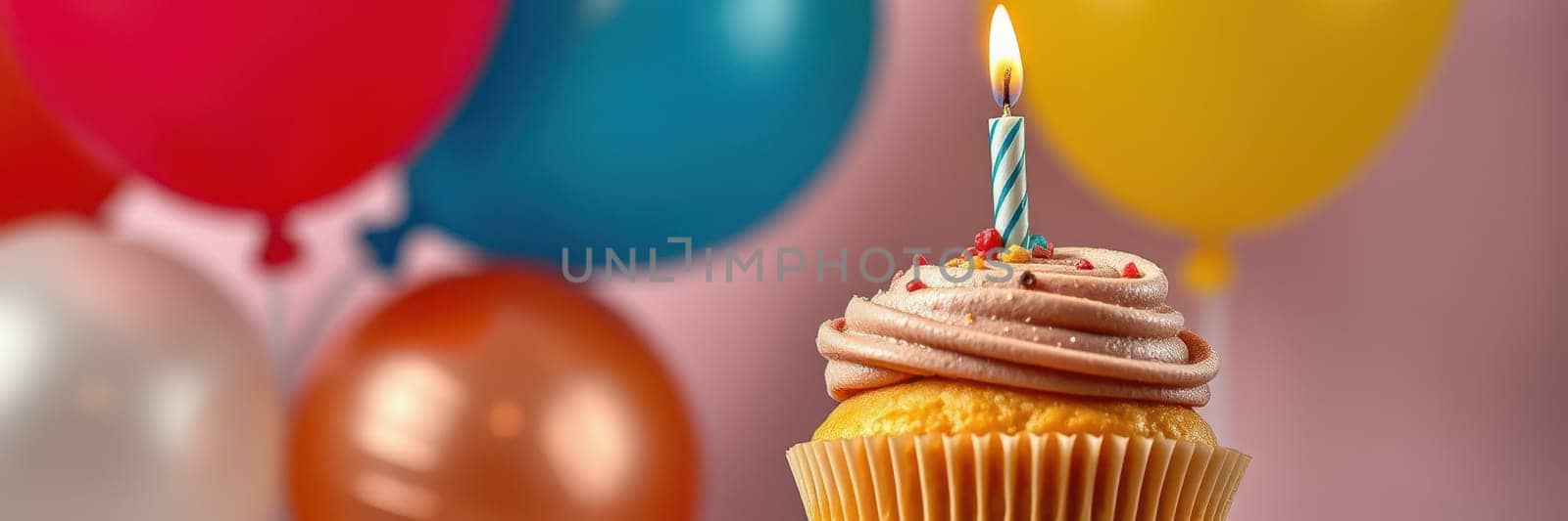 A bright and festive setup features a cupcake adorned with pink frosting and a lit candle, with vibrant balloons in various colors creating a cheerful atmosphere for a celebration.