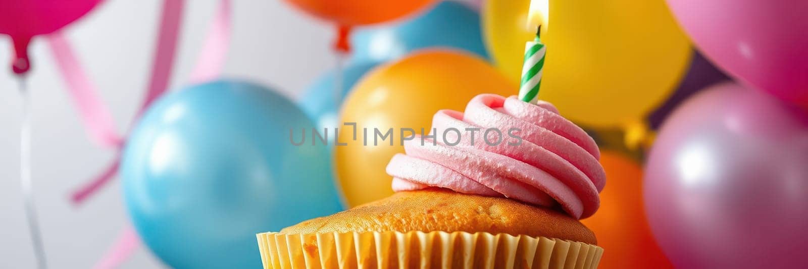 A delicious cupcake topped with pink frosting and a lit candle sits in front of vibrant balloons of various colors, celebrating a joyful birthday occasion.