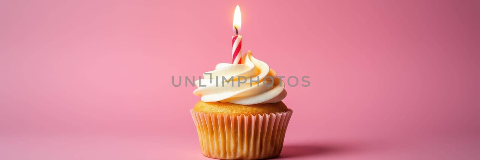 A vanilla cupcake sits on a table adorned with swirls of creamy frosting and a lit candle. The vibrant pink background enhances the festive atmosphere perfect for a birthday.