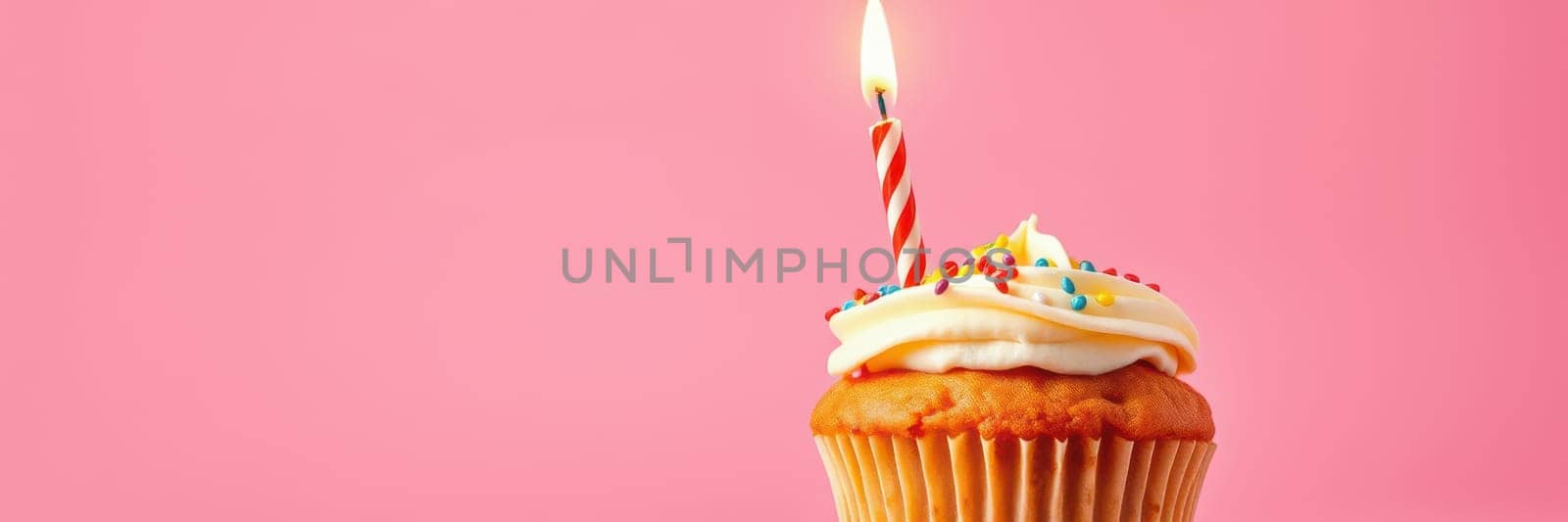 A delightful cupcake topped with creamy frosting and colorful sprinkles features a lit candle on a vibrant pink backdrop, creating a festive atmosphere for celebration.