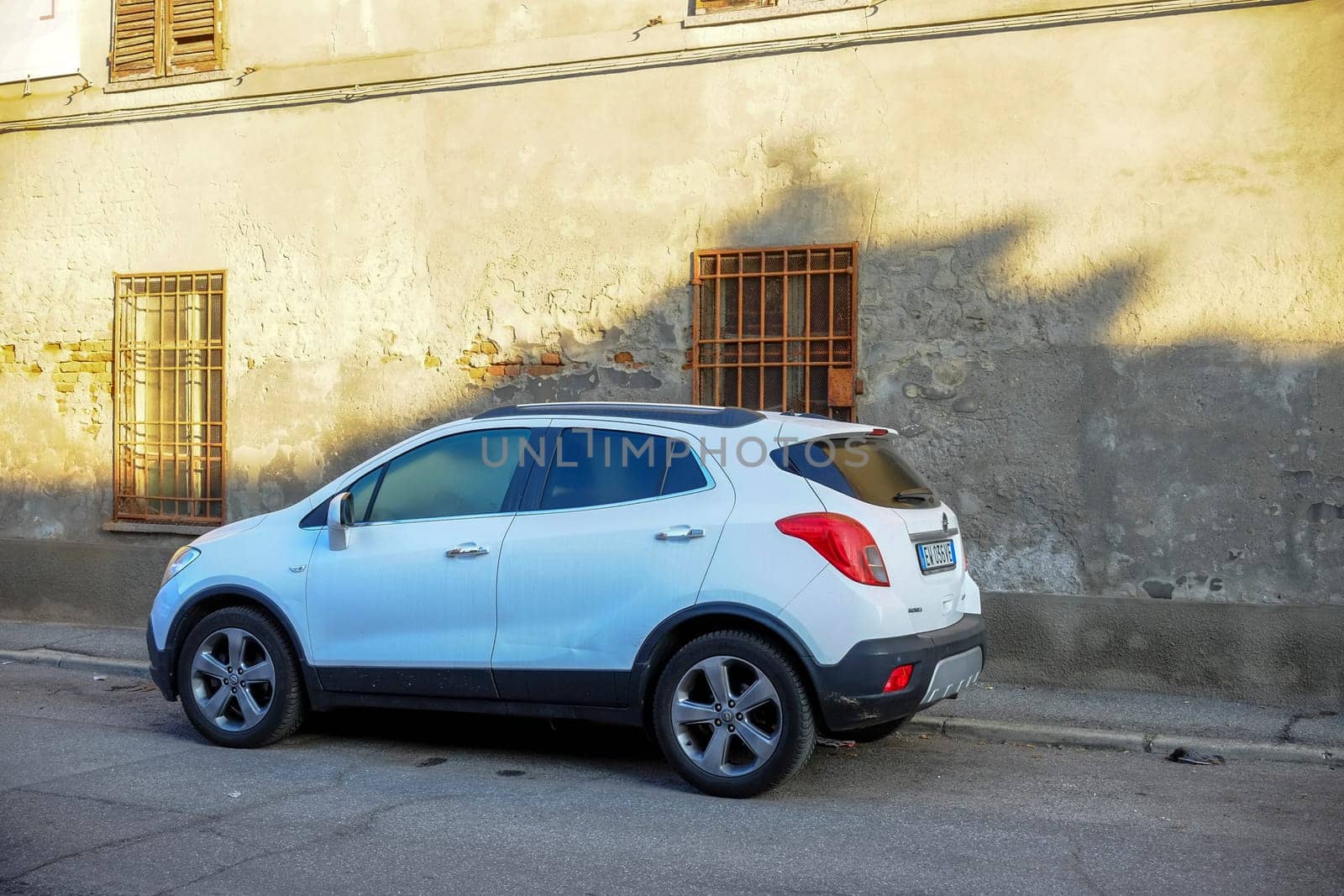 Cremona, Itally - October 6th 2024 A white opel mokka suv is parked on the sidewalk in front of an old, weathered building with barred windows, bathed in the warm glow of late afternoon sunlight