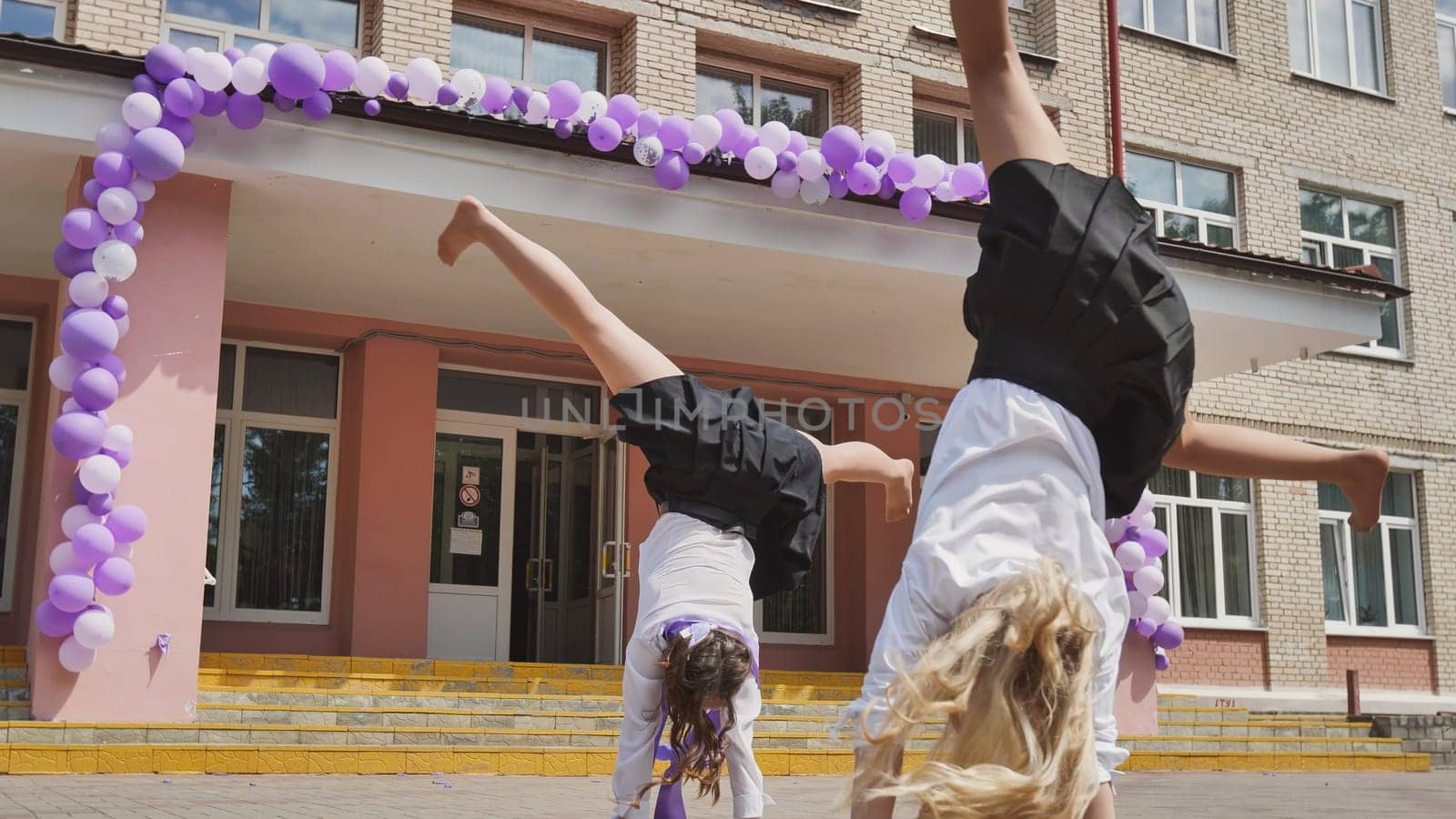 Two schoolgirls performing cartwheels in front of their school on the final day of school, marking the occasion with purple and white balloons