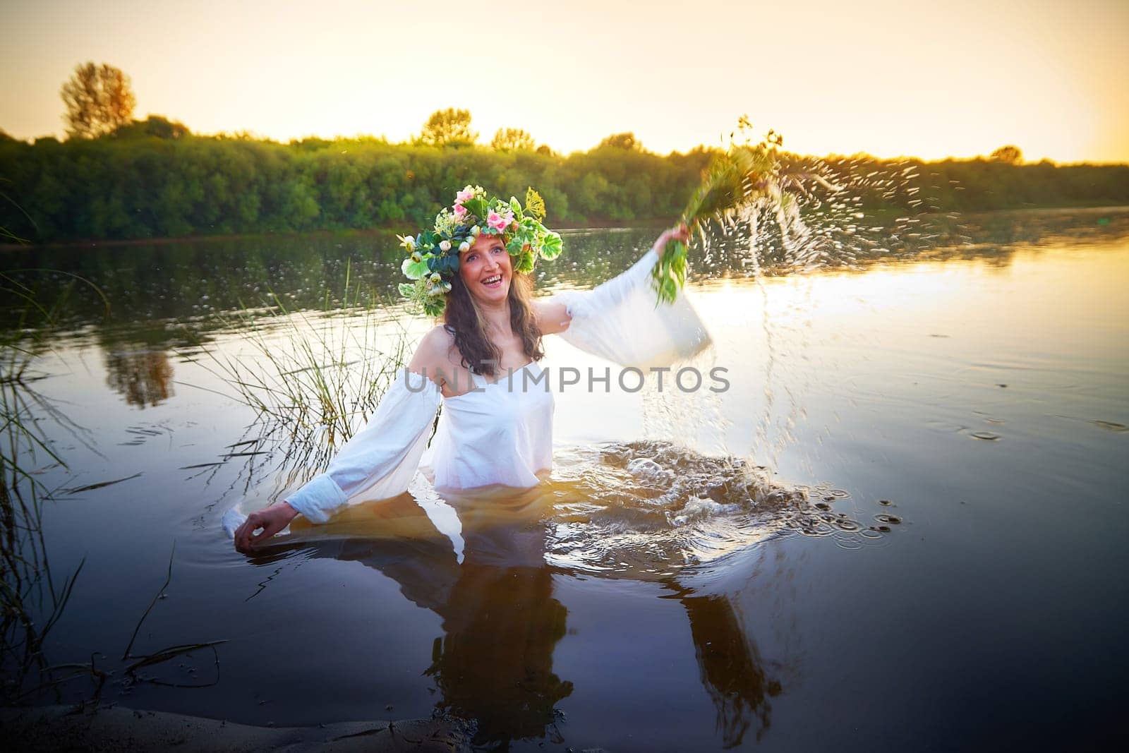 Slavic plump chubby girl in long white dress on the feast of Ivan Kupala with flowers and water in a river or lake on summer evening