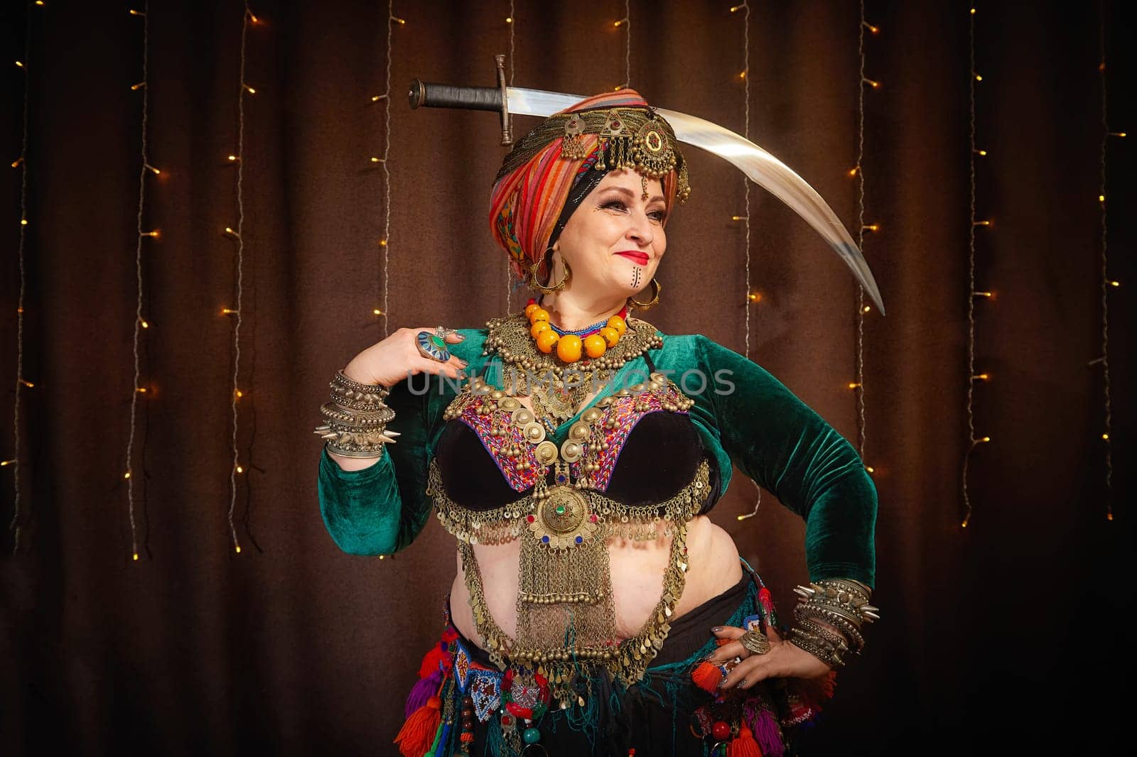 A woman in a colorful headscarf and jewelry poses With a saber in front of wall of colorful tassels