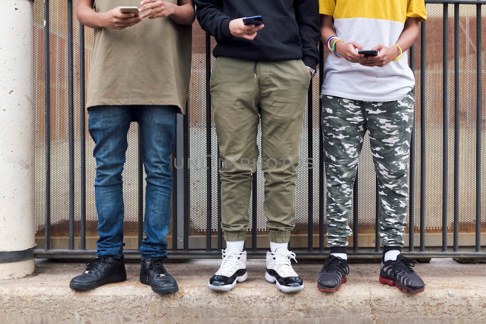 crop of three unrecognizable teen friends browsing Internet on mobile phones standing against metal fence and looking at screen, concept of youth and technology of communication