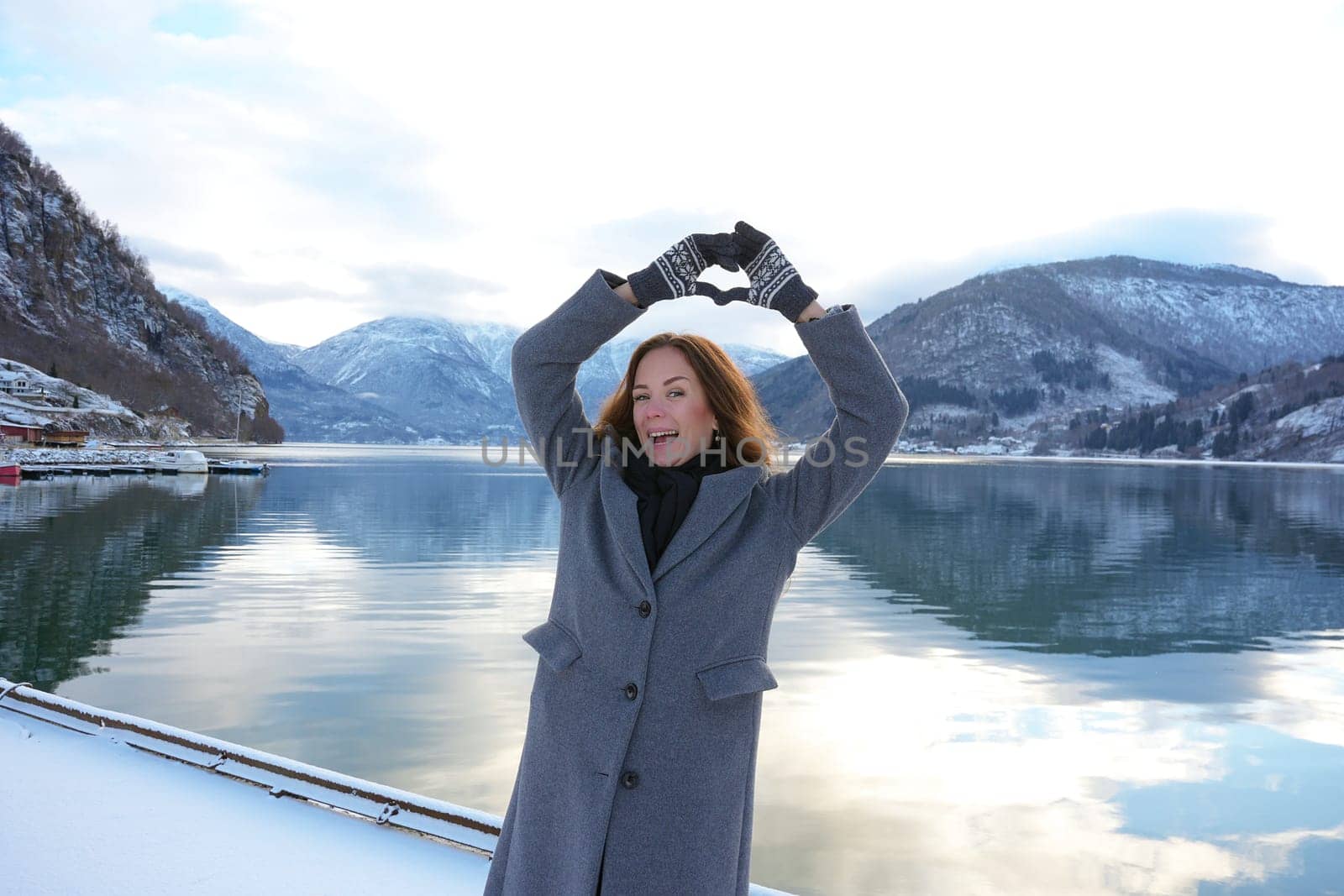 Happy woman in a coat and mittens forms a heart with her hands by a serene snowy fjord and mountains in Gaupne, Norway