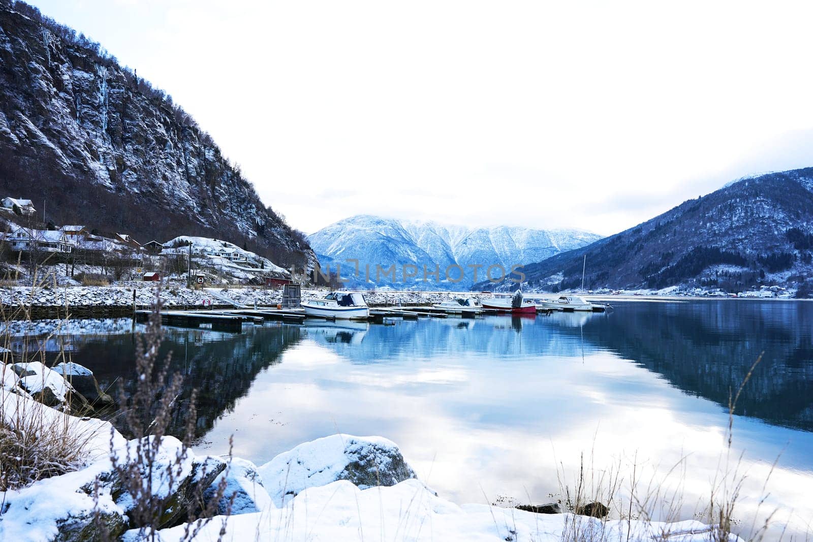 A scenic winter marina in Gaupne, Norway, with snow-covered mountains, calm waters, and boats docked along the fjord's edge. 