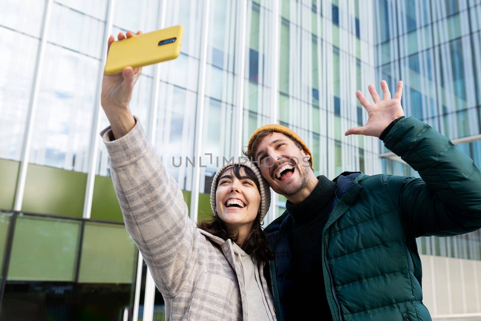 Cheerful man and woman taking a selfie with a yellow smartphone, waving and smiling in front of a modern office building