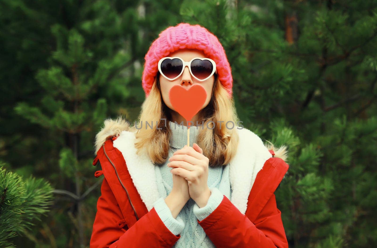 Winter portrait of happy young woman having fun with sweet red heart shaped lollipop wearing pink hat, white glasses against Christmas tree in forest