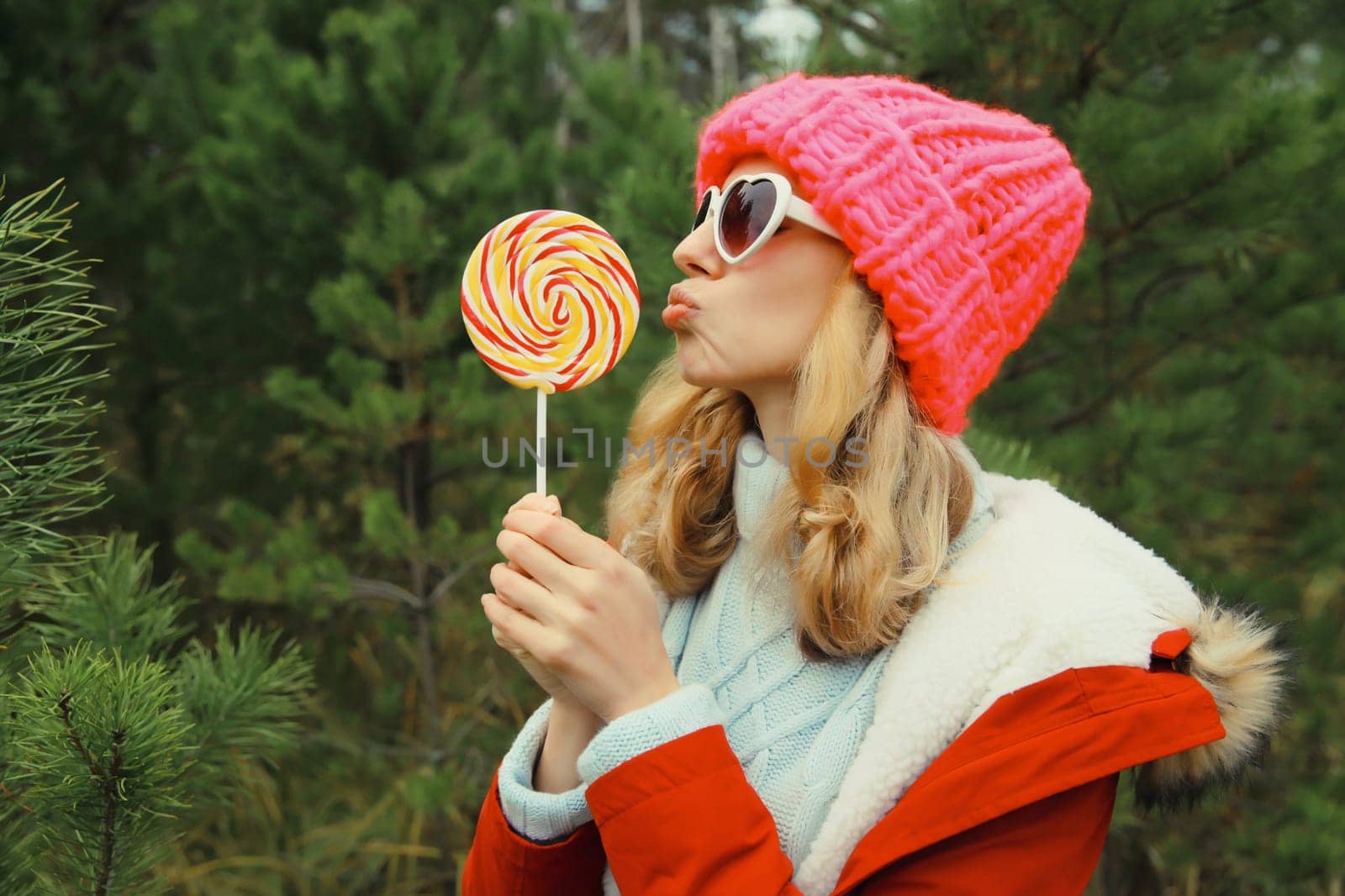 Winter portrait of happy funny smiling young woman having fun with sweet lollipop wearing hat against Christmas tree in forest