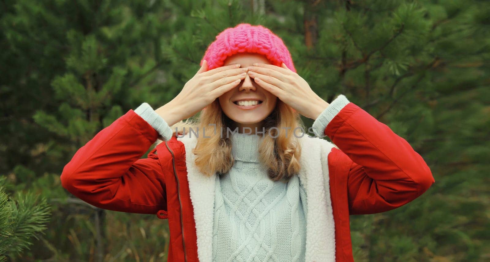 Happy smiling young woman having fun enjoys snow, warm weather in winter park against Christmas tree, joyful girl in hat standing in forest with snowflakes, festive background