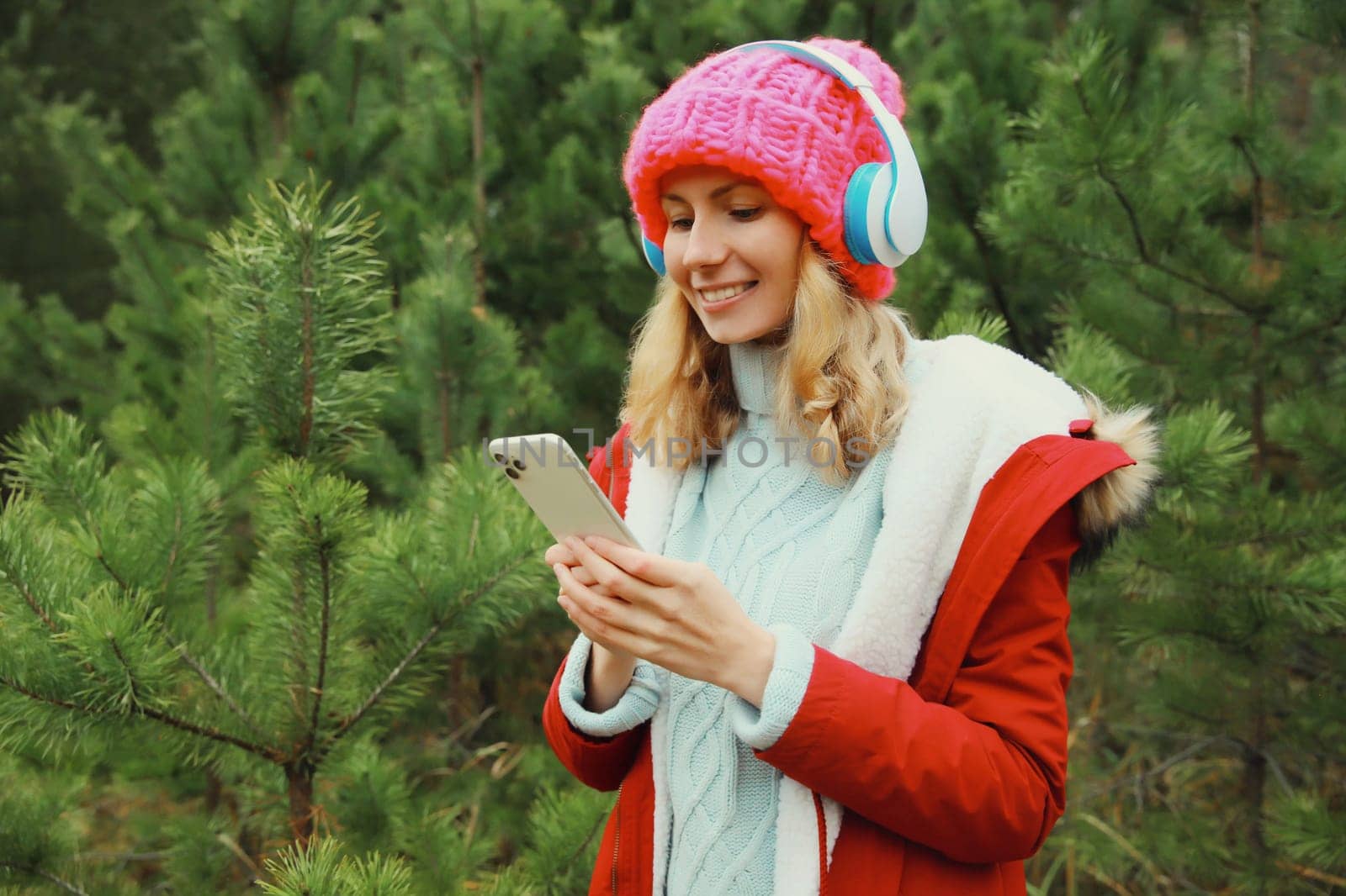 Portrait of happy modern young woman with phone enjoying listening to music in headphones in hat, jacket in winter forest against Christmas tree background