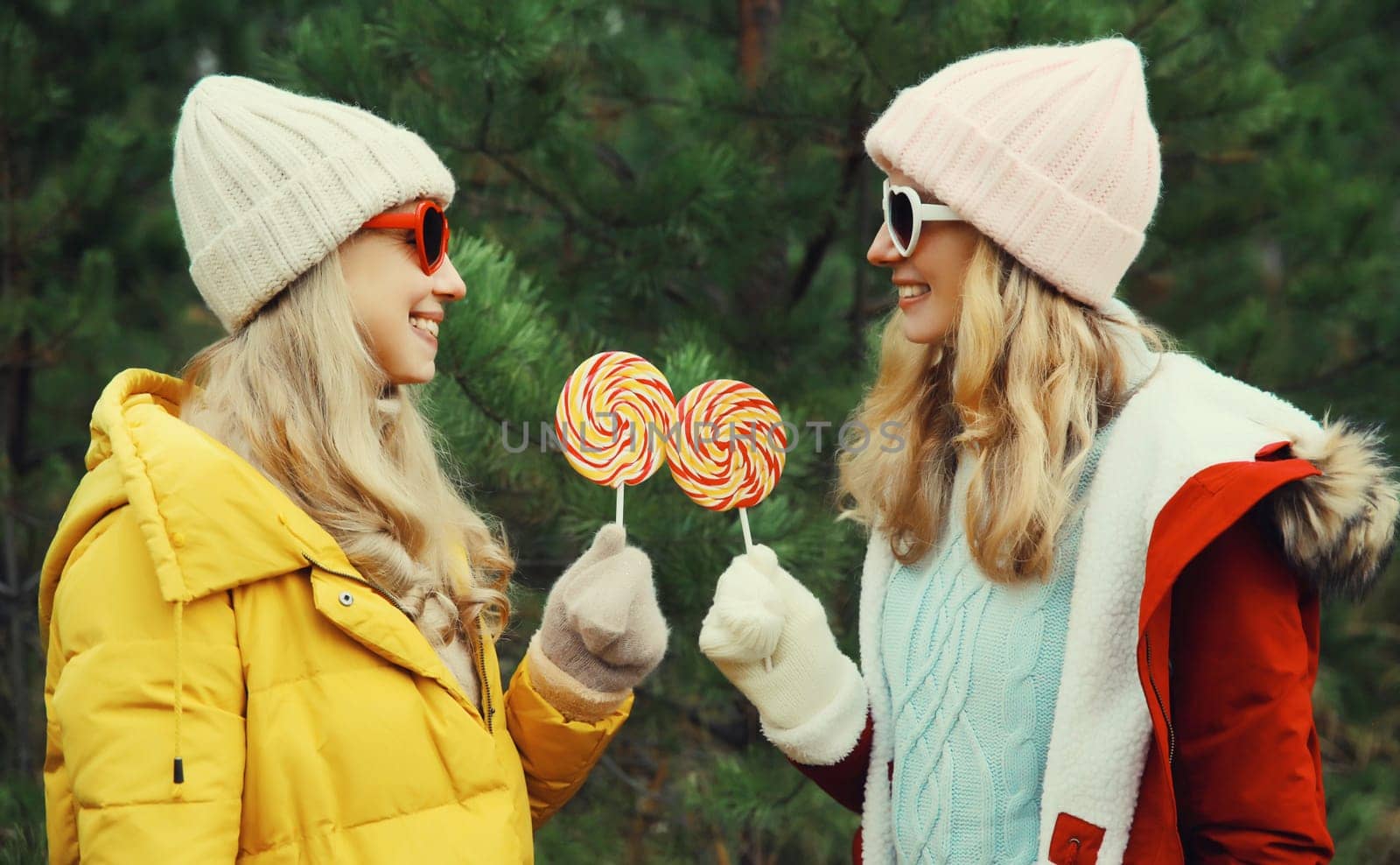 Happy smiling women friends having fun, celebration, joyful girlfriends together on Christmas tree background in winter forest, wearing hat, colorful jacket outdoors