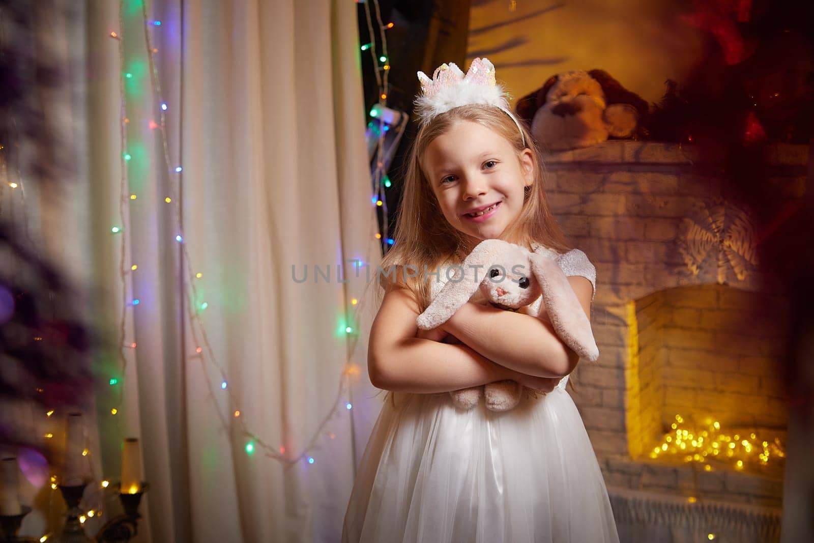 Little girl smiles while embracing her plush bunny in a warmly lit, decorated room filled with holiday cheer and lights.