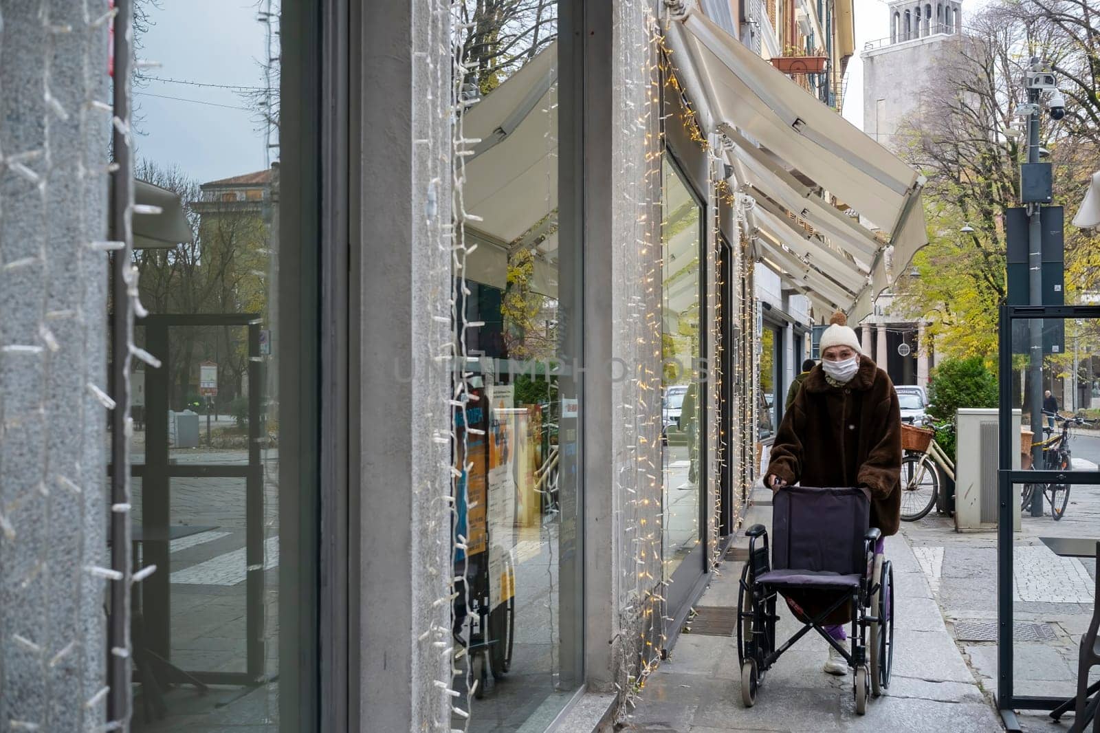 Cremona, Italy - November 20th 2024 Senior woman wearing a surgical mask and pushing a wheelchair on a decorated street in cremona, italy