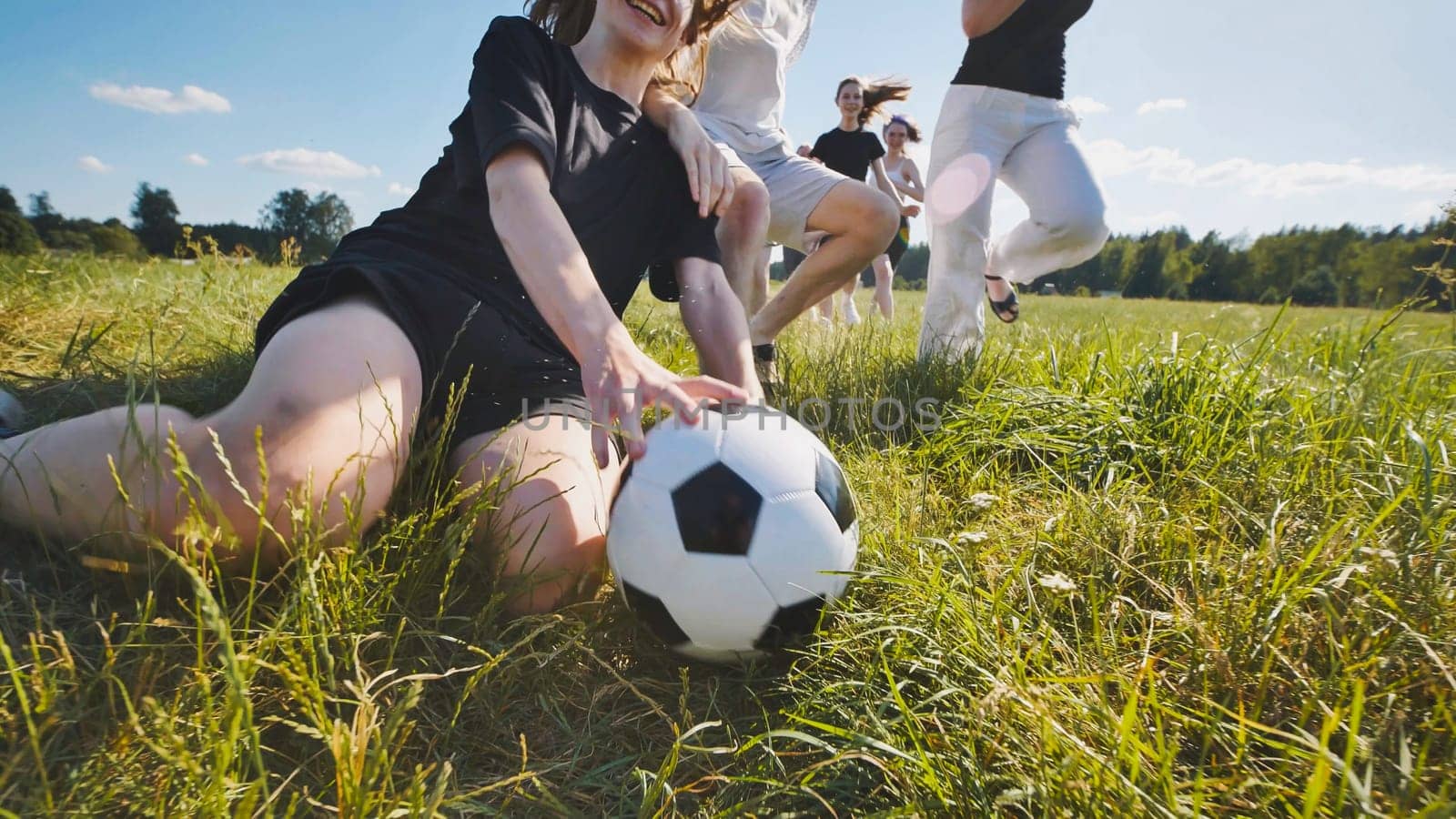 Group of young adults is running after a soccer ball on a grass field on a sunny summer day