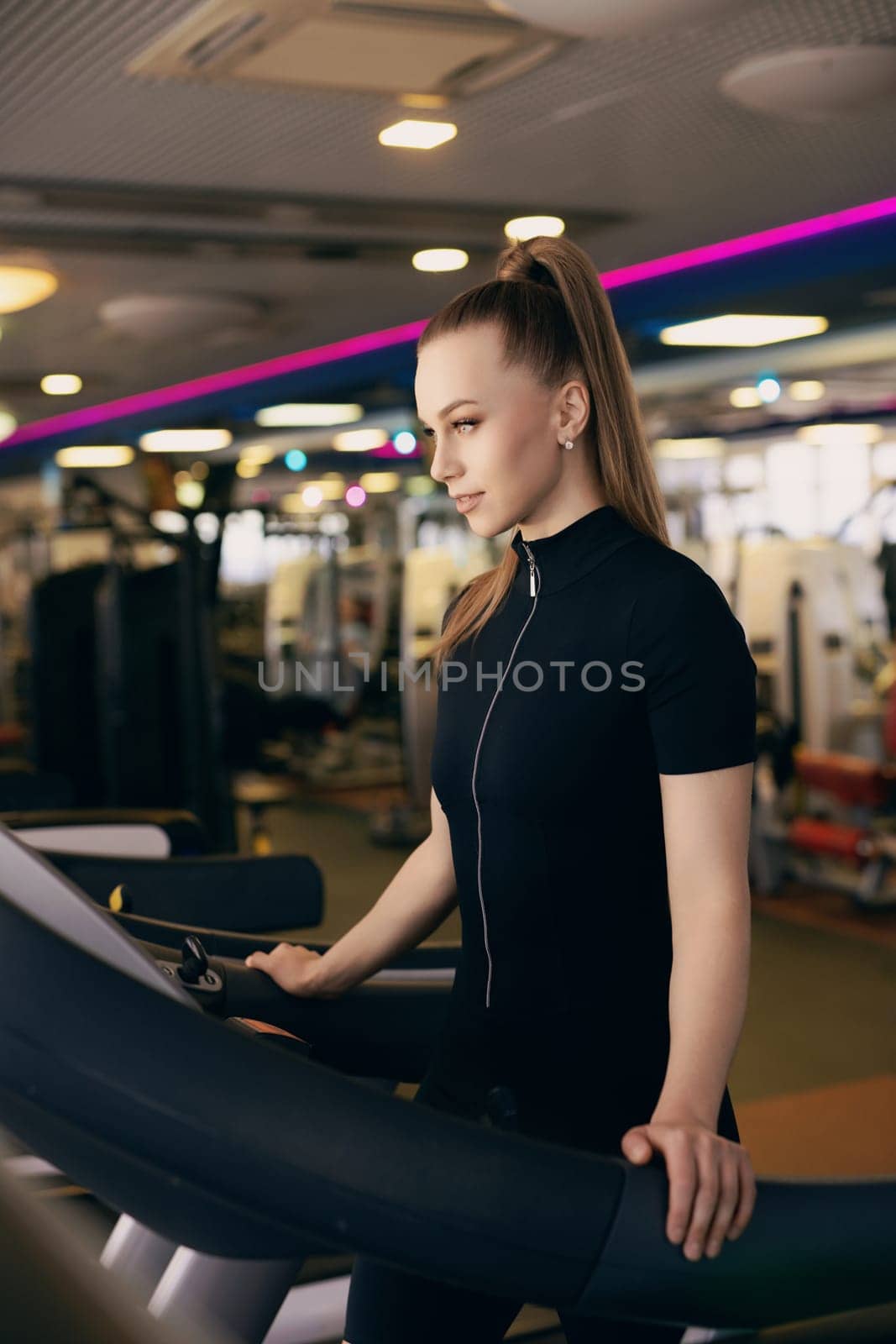 A young woman with a ponytail exercises on a treadmill in a bright, modern gym filled with fitness equipment. The atmosphere is energetic and motivating, showcasing a health-focused environment.