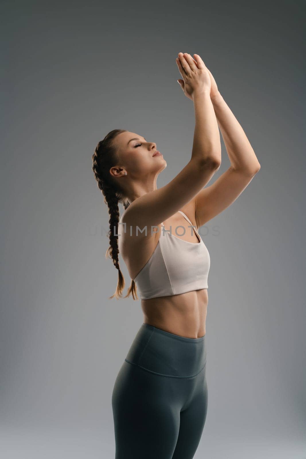 A woman in a sports bra and yoga pants performs a pose with her hands together, eyes closed, in a peaceful studio. Soft lighting enhances the tranquil atmosphere of her practice.