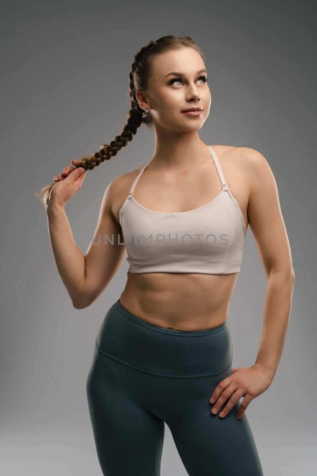 A young woman stands with a confident expression, wearing a sports bra and high-waisted leggings. She has a braided hairstyle and maintains an athletic pose, showcasing her fit physique.