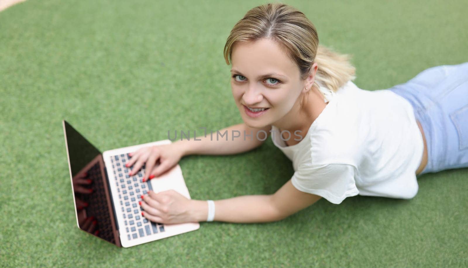 Young woman freelancer with laptop lies on green cover, face close-up, top view. Work while resting, business
