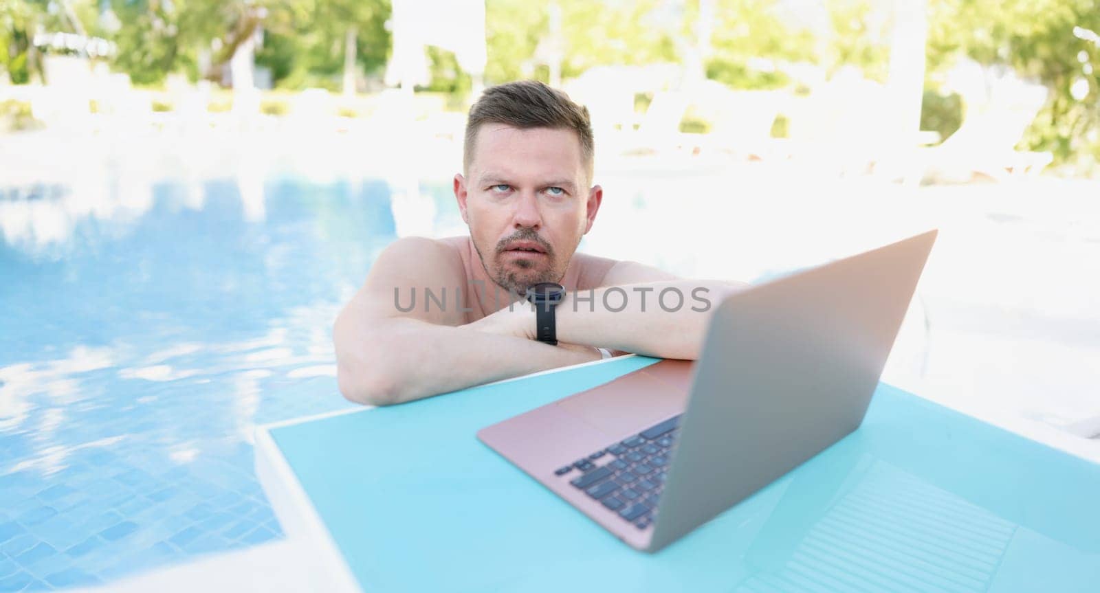Annoyed man working on a laptop from the pool, close-up. Unexpected remote work while on vacation, distraction from rest