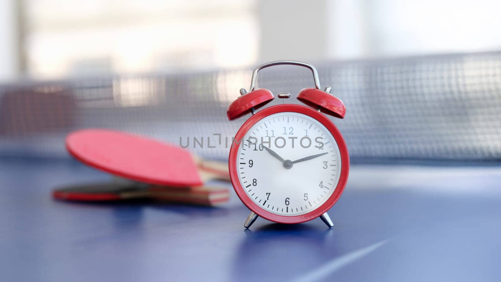 Red alarm clock on a tennis table, close-up, blurry. Office ping pong during the break. Sports recreation for employees