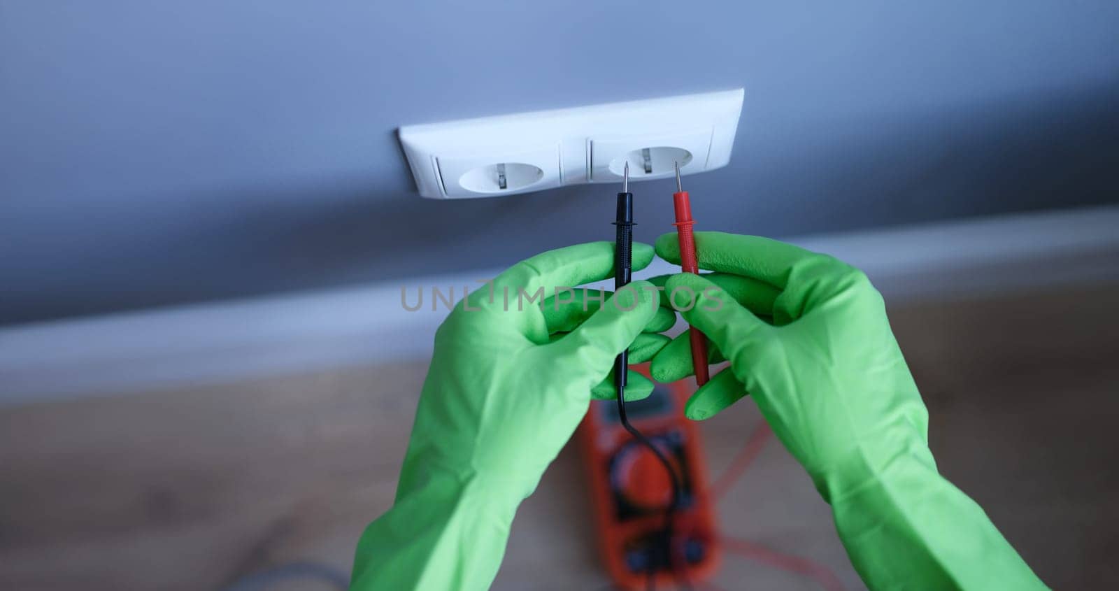 Gloved hands are holding a digital multimeter near the socket, close-up. Test device. AC and DC current measurement
