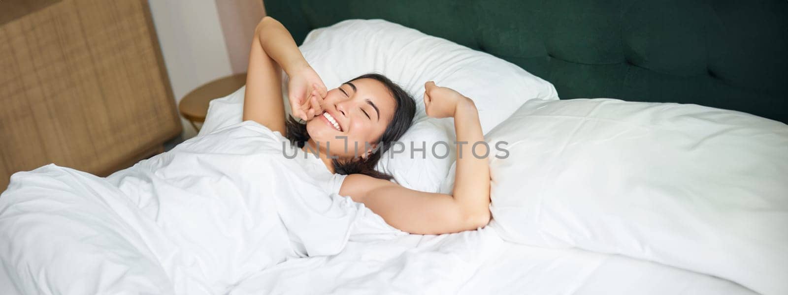 Happy mornings. Beautiful asian girl wakes up in a hotel suit, lying in white bed, stretching her arms and smiling happily.