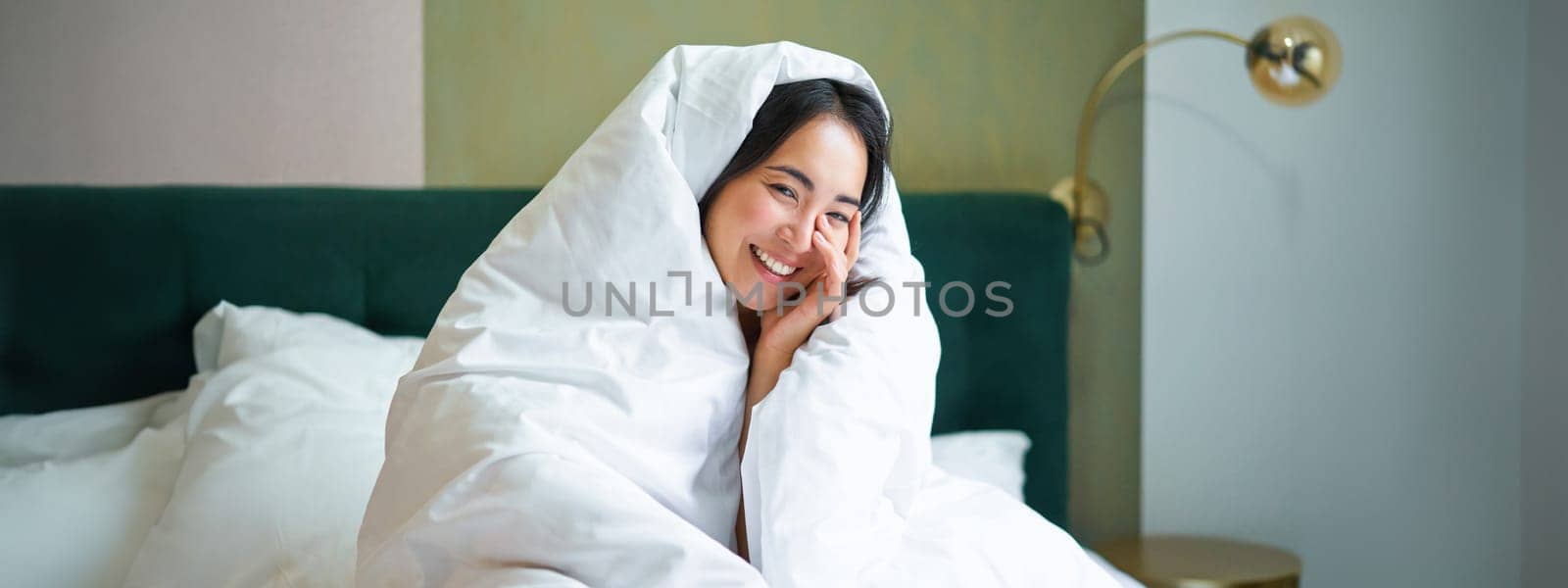 Beautiful asian woman sitting on bed, covered in blankets and duvet, laughing and smiling, enjoying weekend in bedroom by Benzoix