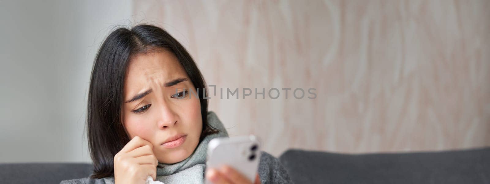 Ill korean woman sitting at home, sneezing, caught cold, reading news on mobile phone, freezing at her home because of heating problems by Benzoix