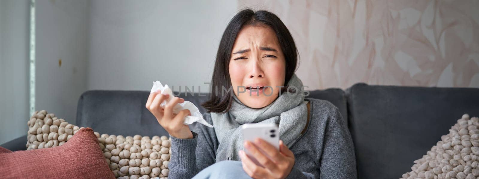 Asian girl with influenza, sits at home, cries and looks upset, holds smartphone, feels unwell and gloomy, wearing warm clothes.