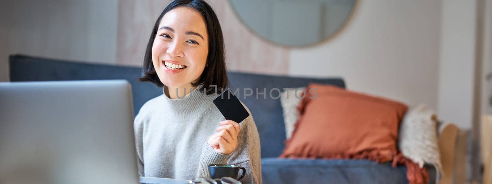 Happy smiling girl with credit card, paying her bills online on computer, doing shopping on her laptop, sitting at home.