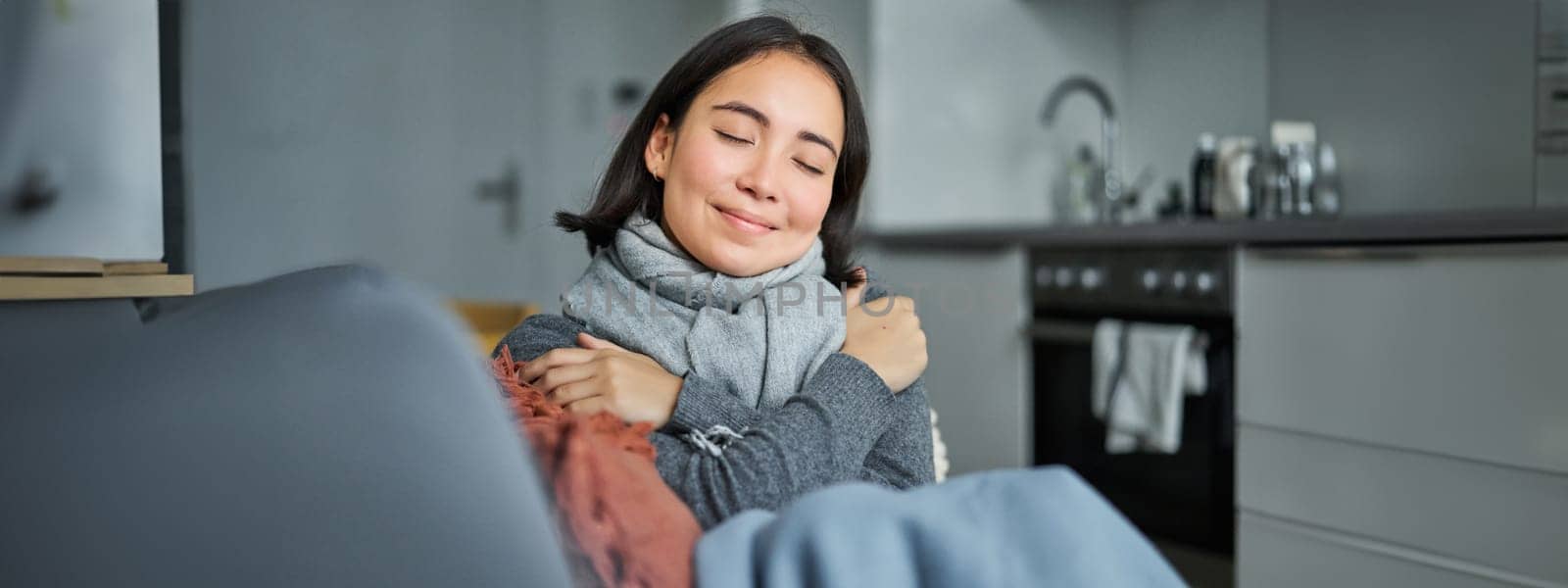 Portrait of smiling asian woman feels warmth, hugs herself with pleased face expression, warming up in clothes and scarf at home without heating.