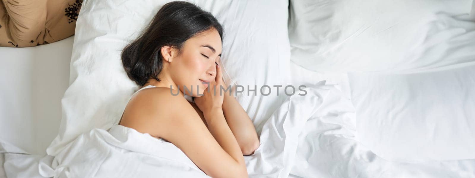 Top view image of asian woman sleeping alone in king size bed on white pillows. Young girl lying in her bedroom with eyes closed, morning sunlight shines in room.