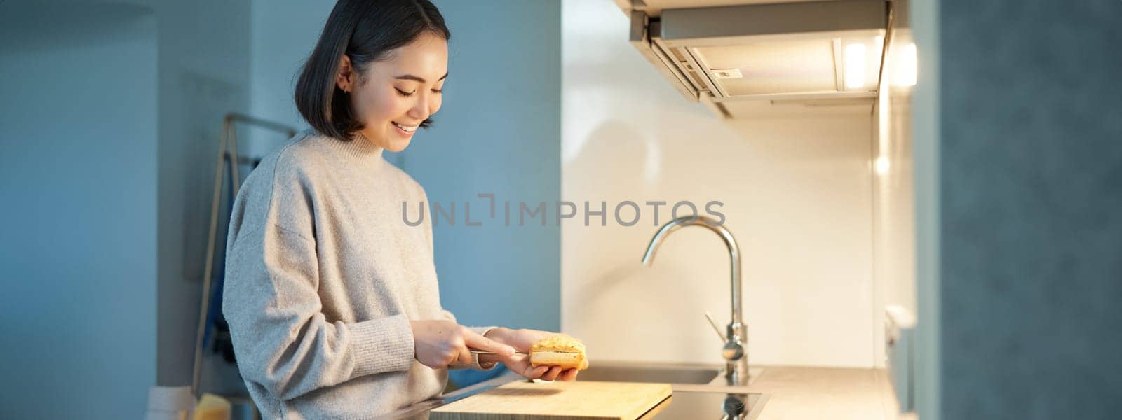 Portrait of smiling young asian woman standing on kitchen and making a sandwitch, cooking for herself.