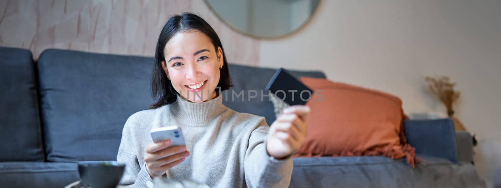Smiling cute asian woman using credit card and smartphone, paying bills online, holding mobile phone, looking at camera.