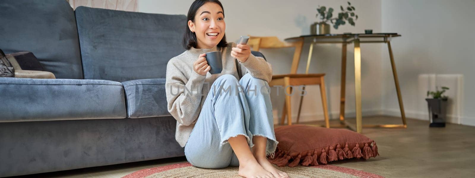 Portrait of girl watching television at home, sits on floor near sofa, holds remote and changes channels.