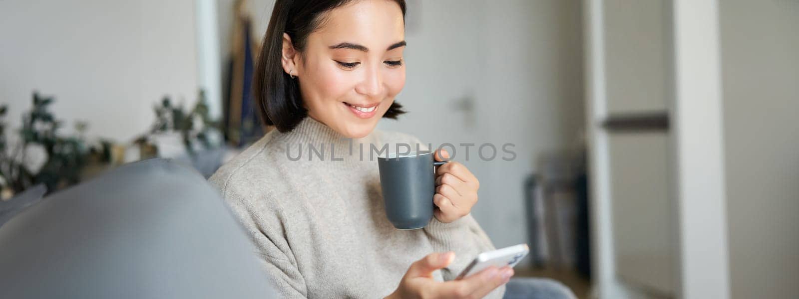 Portrait of smiling asian girl checking her news feed on smartphone and drinking coffee, sitting on sofa at home, browsing on mobile phone, reading.