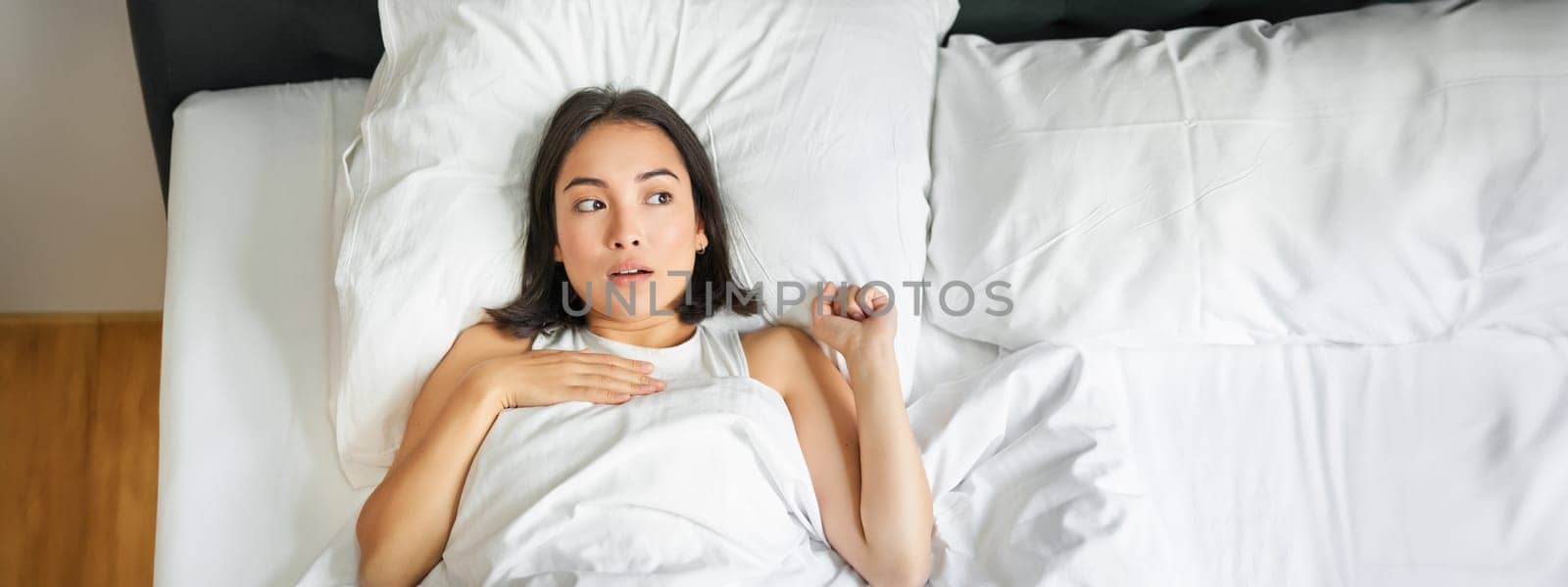 Portrait of asian woman lying in bed with shocked face, looking startled and upset, gasping from smth, staying alone in her bedroom.