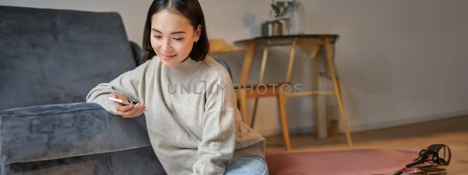 Vertical shot of young woman in cozy home working on laptop, using smartphone and drinking coffee, sitting on floor near sofa by Benzoix